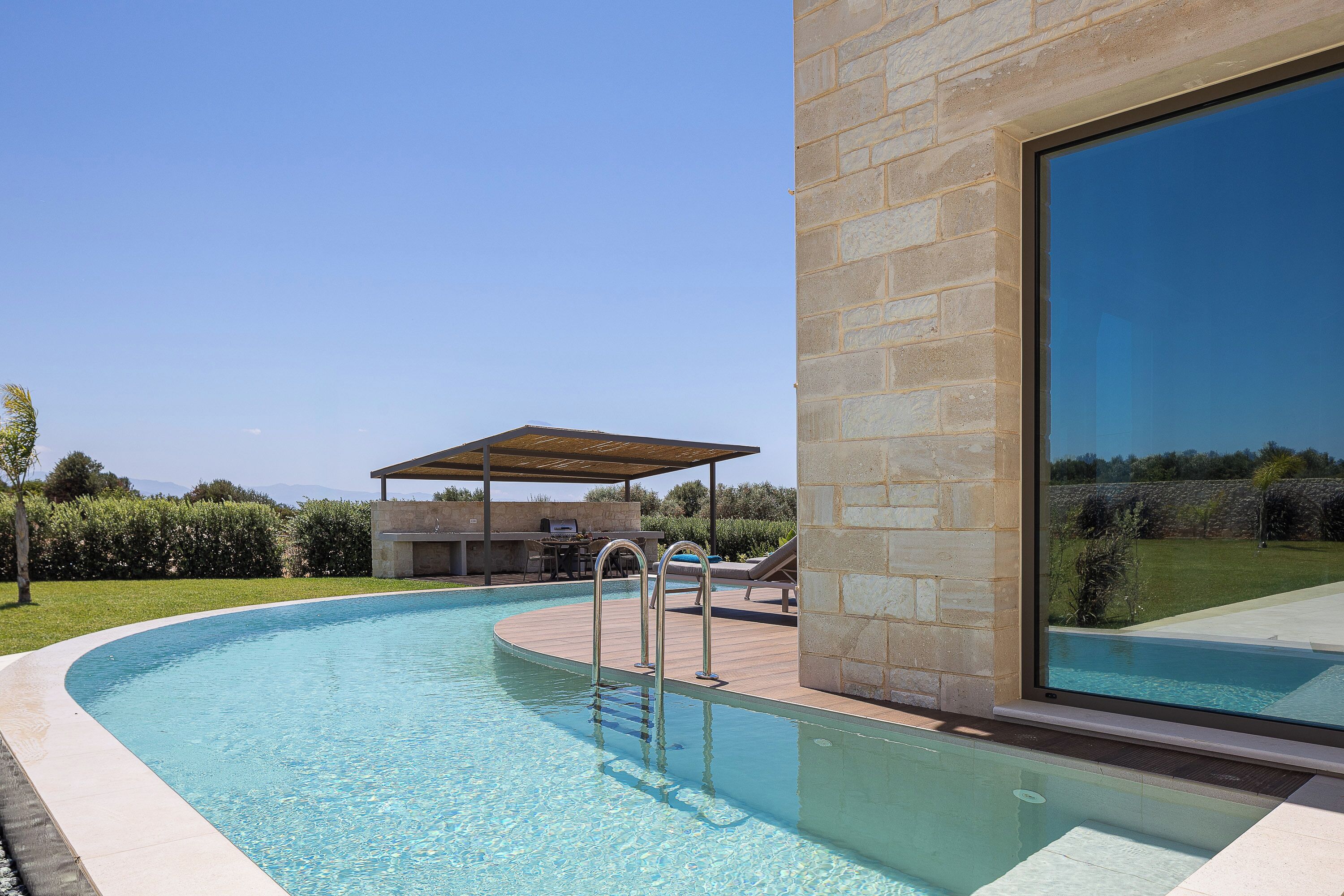 A swimming pool with a wooden deck, a covered barbecue area, and a stone wall.