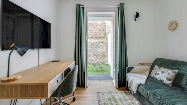 Une pièce comprenant un bureau en bois, une chaise, un canapé vert, un téléviseur fixé au mur et une vue sur l’extérieur par une porte munie de rideaux.