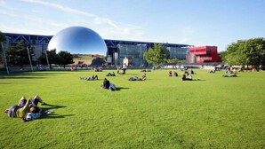 Un espace verdoyant et dégagé où des personnes se détendent, une grande structure sphérique et, au loin, des bâtiments modernes.