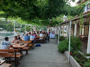 An outdoor dining area with wooden tables and benches, surrounded by greenery and overlooking a body of water. - Apartments Franka Saplunara-triple Room With Terrace and Sea View No1 (Mljet)