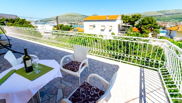 A terrace with a table set for two, a bottle of wine, and a glass, overlooking a coastal town with buildings and hills. - Apartments Ivana - Studio With Balcony and Sea View (Trogir)