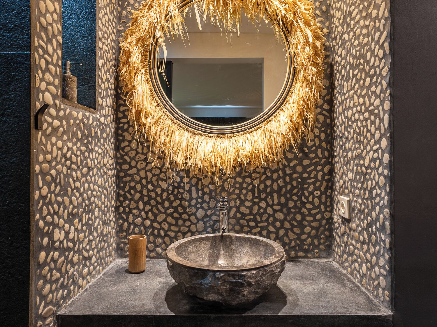 A bathroom with a stone wall, a round mirror with a straw wreath, a stone sink, and a wooden cup.