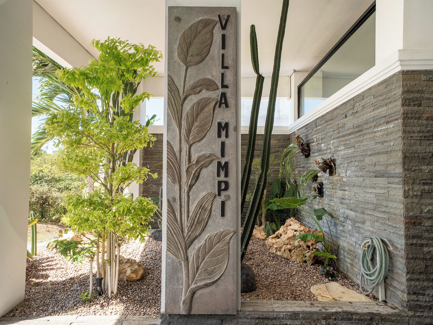 A stone pillar with the inscription 'VILLA MIMPI' in a courtyard with plants and a stone wall.