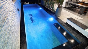Ein Schwimmbecken mit dem Schriftzug 'MIMPI' auf der Wasseroberfläche, umgeben von einer modernen Außenanlage mit Pflanzen und einer Bar.