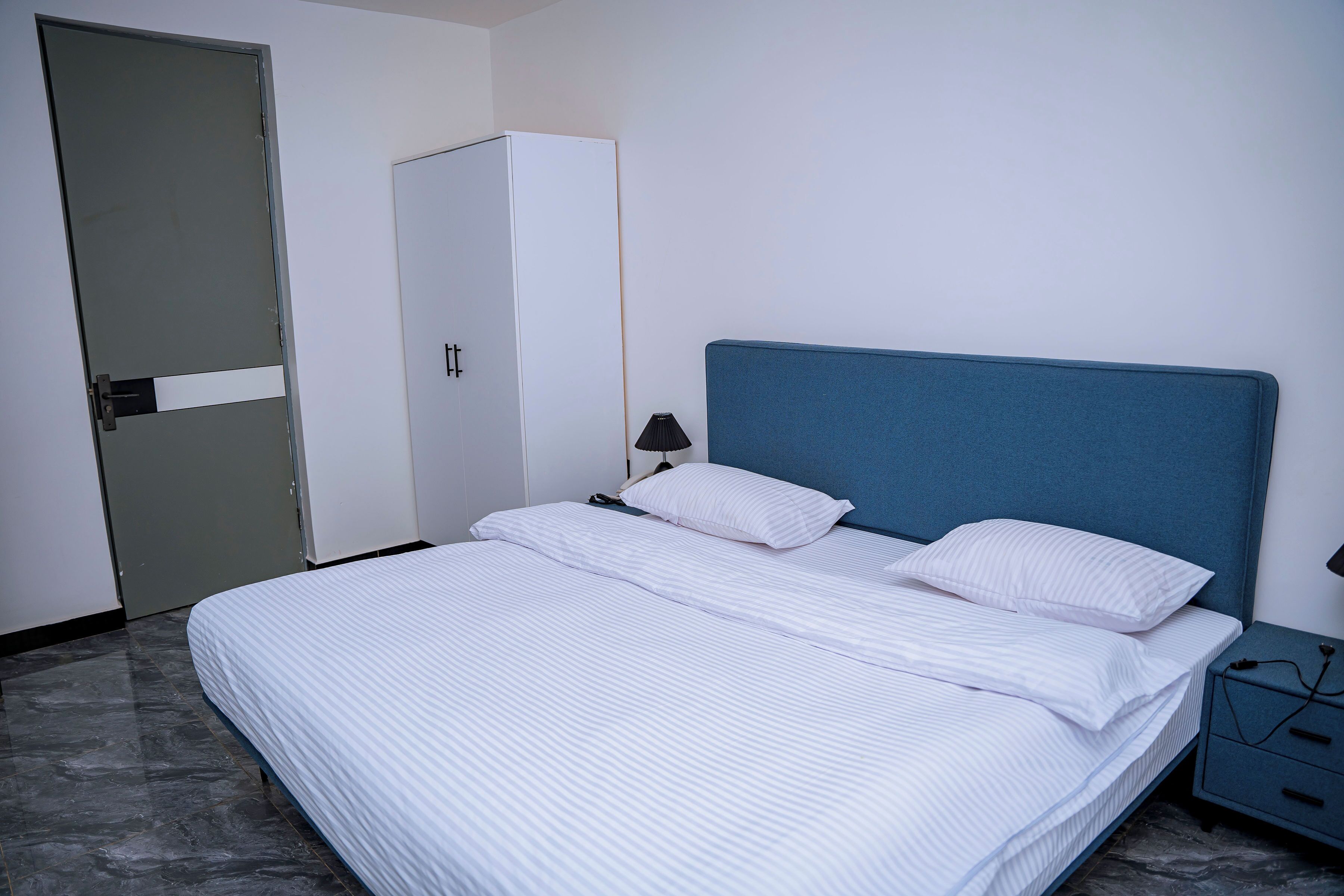 A neatly made bed with white bedding, a blue headboard, and a bedside table with a lamp and charger.