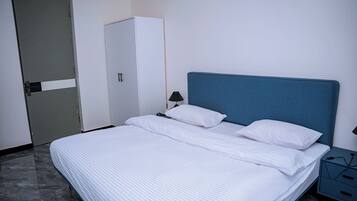 Cama bem arrumada com roupa de cama branca, cabeceira azul e mesa de cabeceira com abajur e carregador.