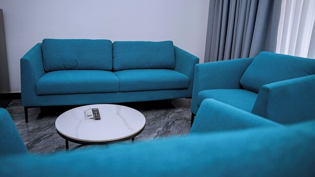 Una habitación con sofá azul y sillones, una pequeña mesa redonda con un control remoto y una ventana con cortinas.