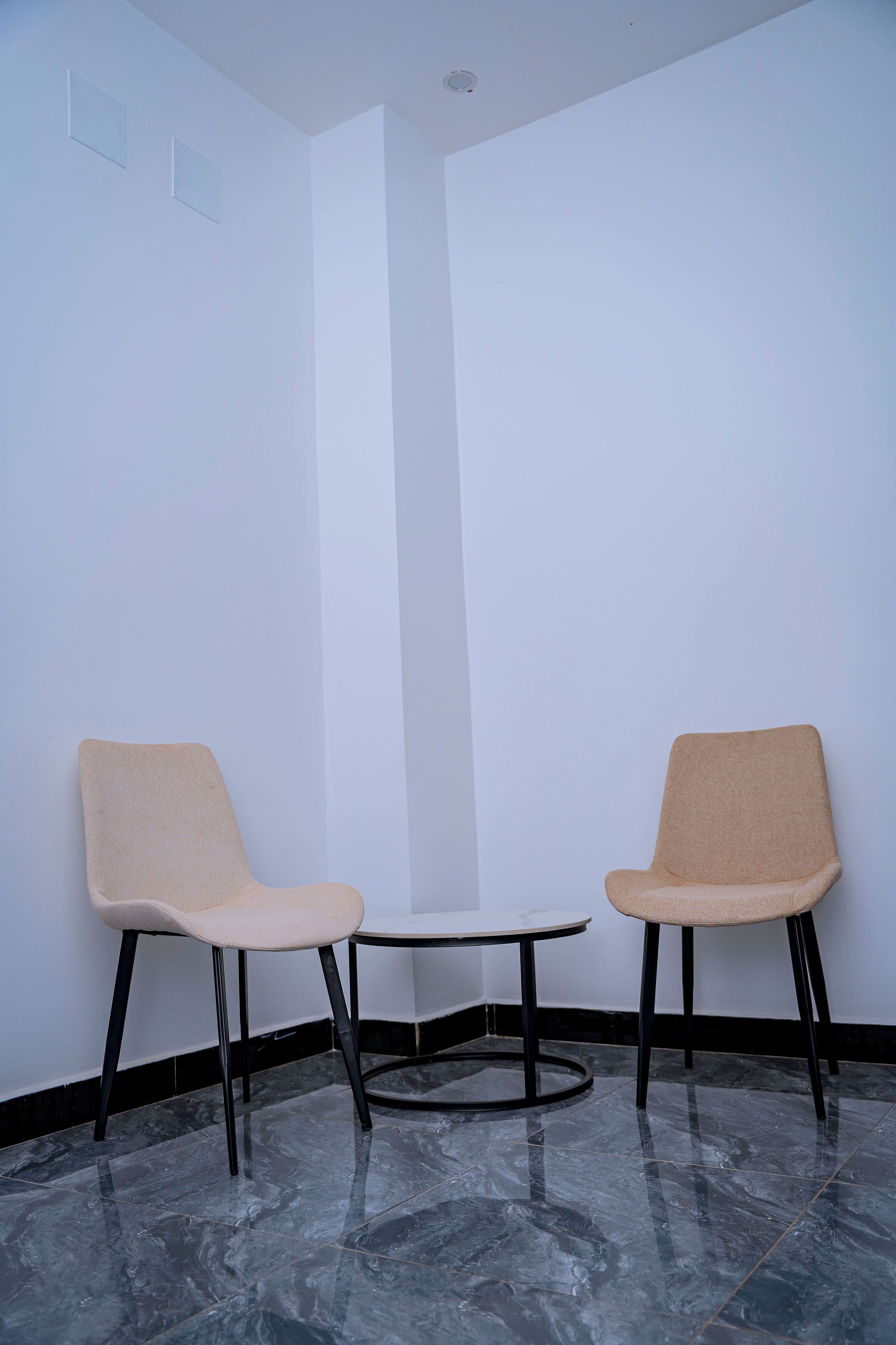 A corner room with two chairs and a small table, white walls, and a marble floor.