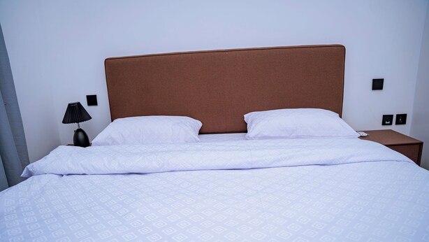 A neatly made bed with a patterned bedspread and two pillows, a bedside lamp, and a nightstand with a lamp. - Ink & S Apartment Hotel (Kigali)