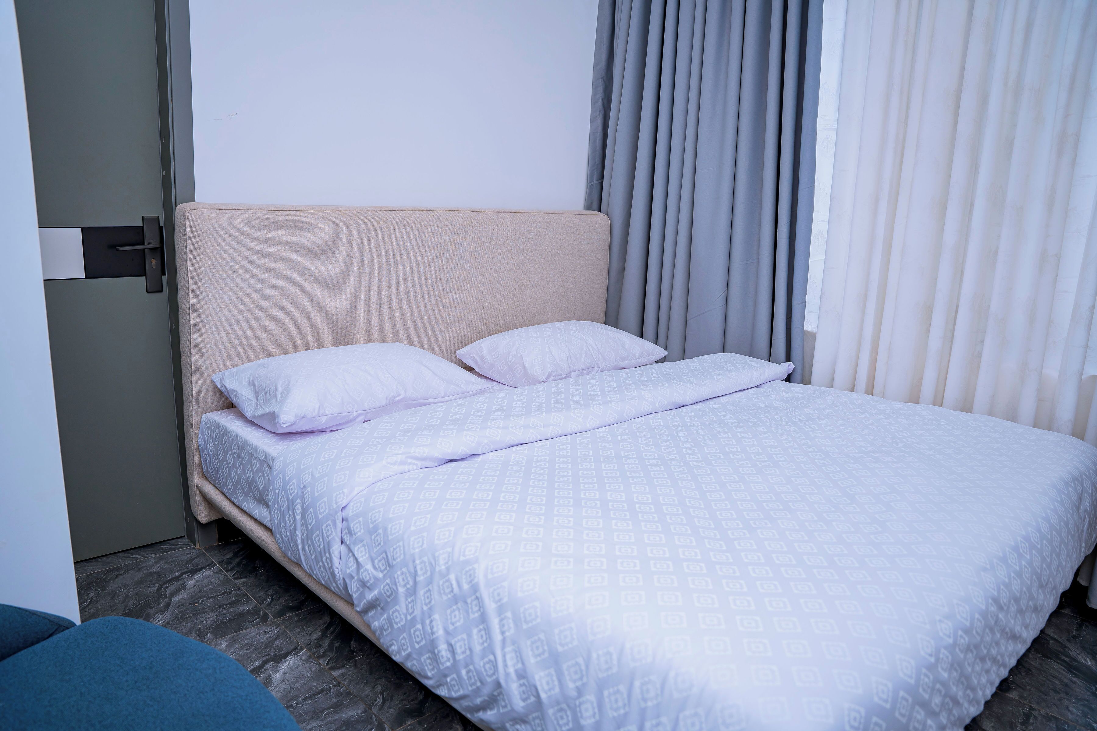 A hotel room with a bed, a headboard, a window with curtains, and a door.