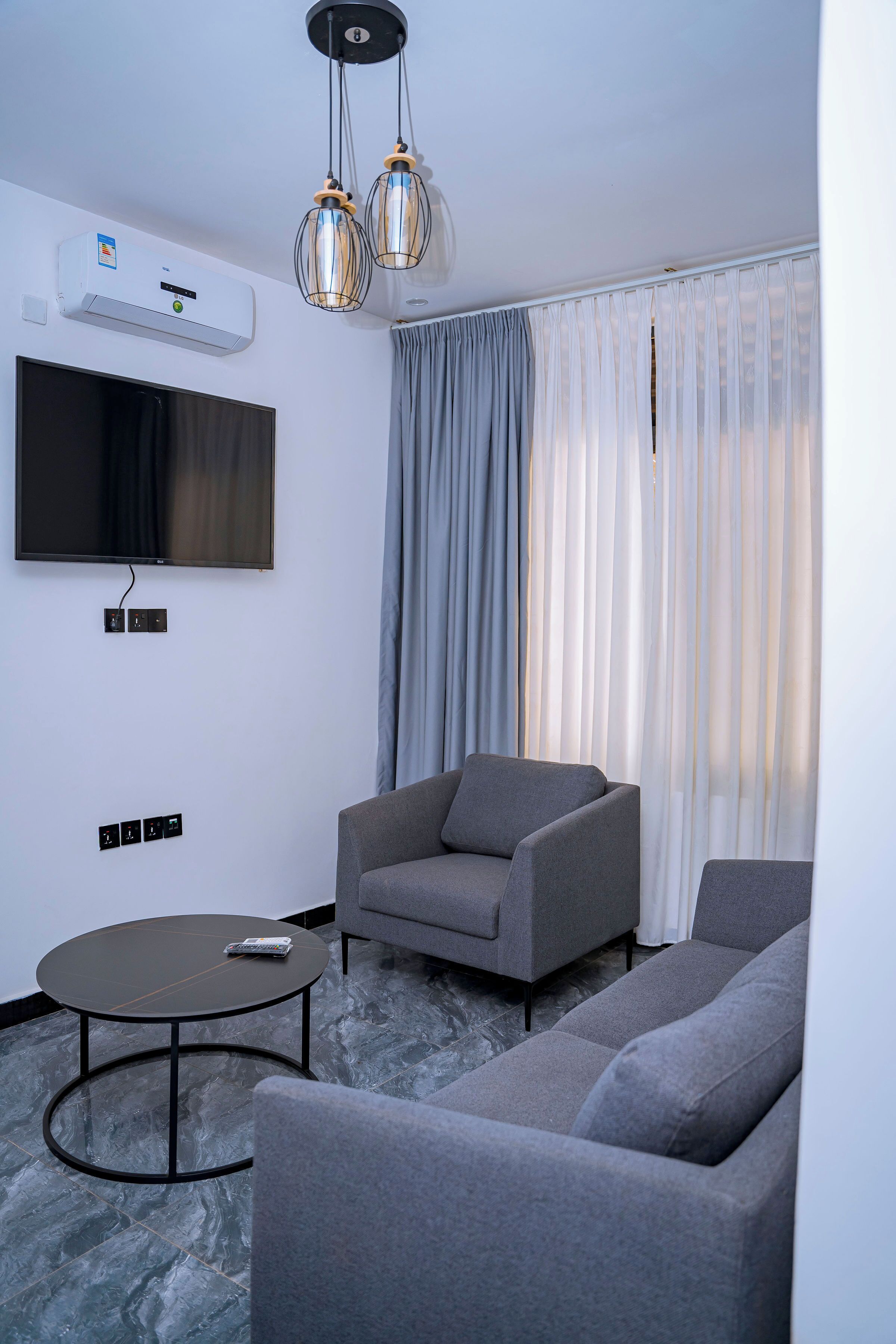A modern living room with a flat-screen TV, grey sofas, a round coffee table, and a ceiling-mounted air conditioning unit.