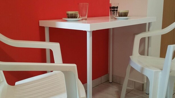 Ein kleiner, weißer Tisch mit einem Glas und zwei Tassen an einer roten Wand, begleitet von weißen Stühlen.