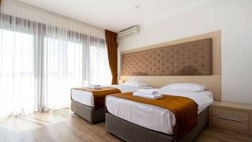 A hotel room with two beds, a large window with curtains, a wall-mounted air conditioner, and a bedside table.