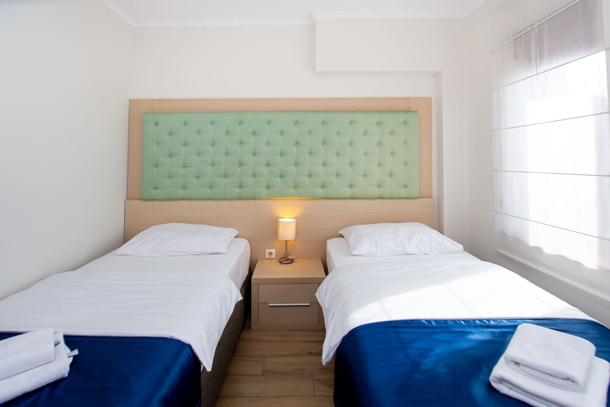 Hotellihuone, jossa on kaksi sänkyä, yöpöytä lampun kanssa ja ikkuna, jossa on sälekaihtimet.