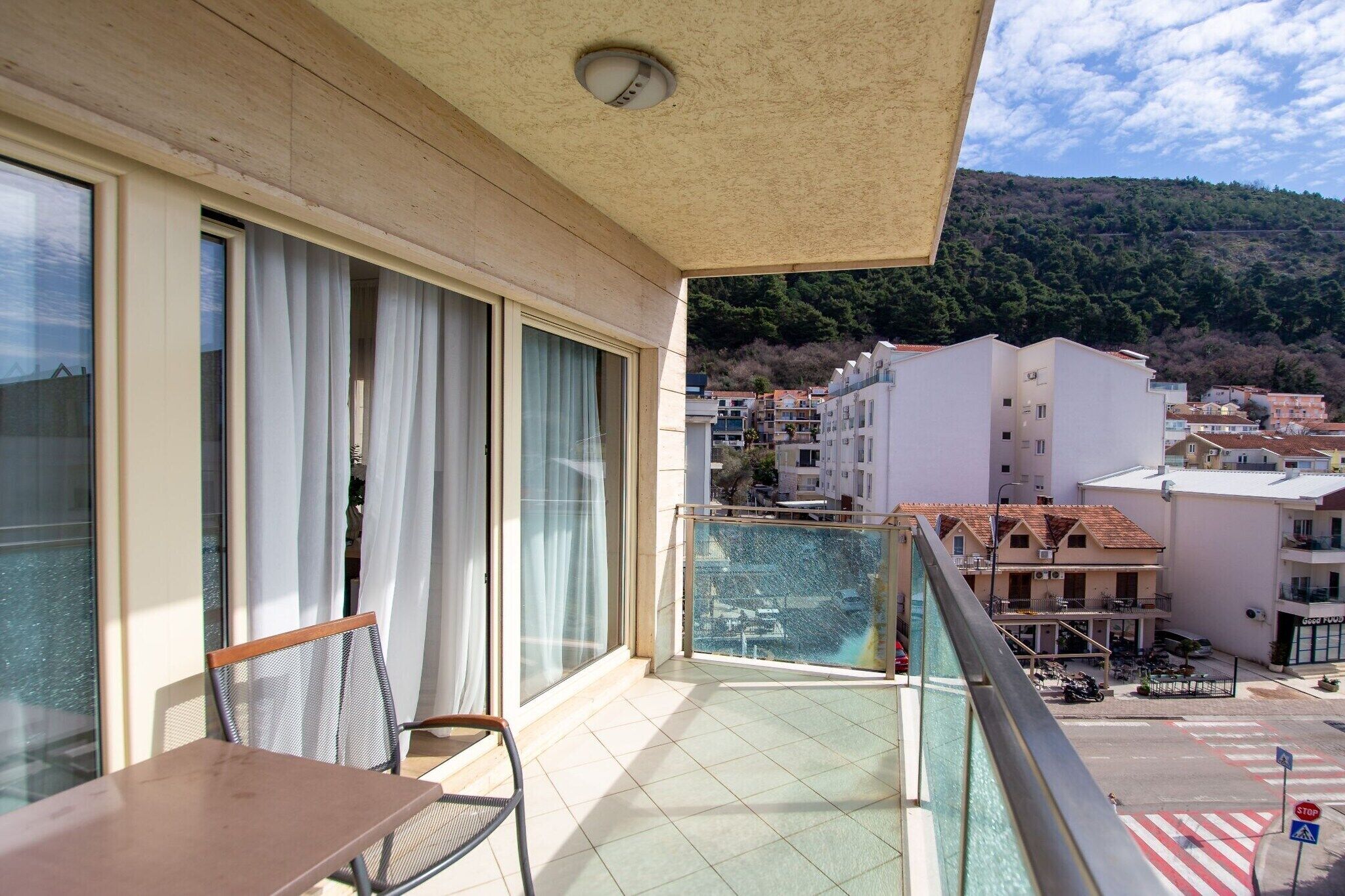 A balcony with a table and chair, overlooking a residential area with buildings and a hilly landscape.
