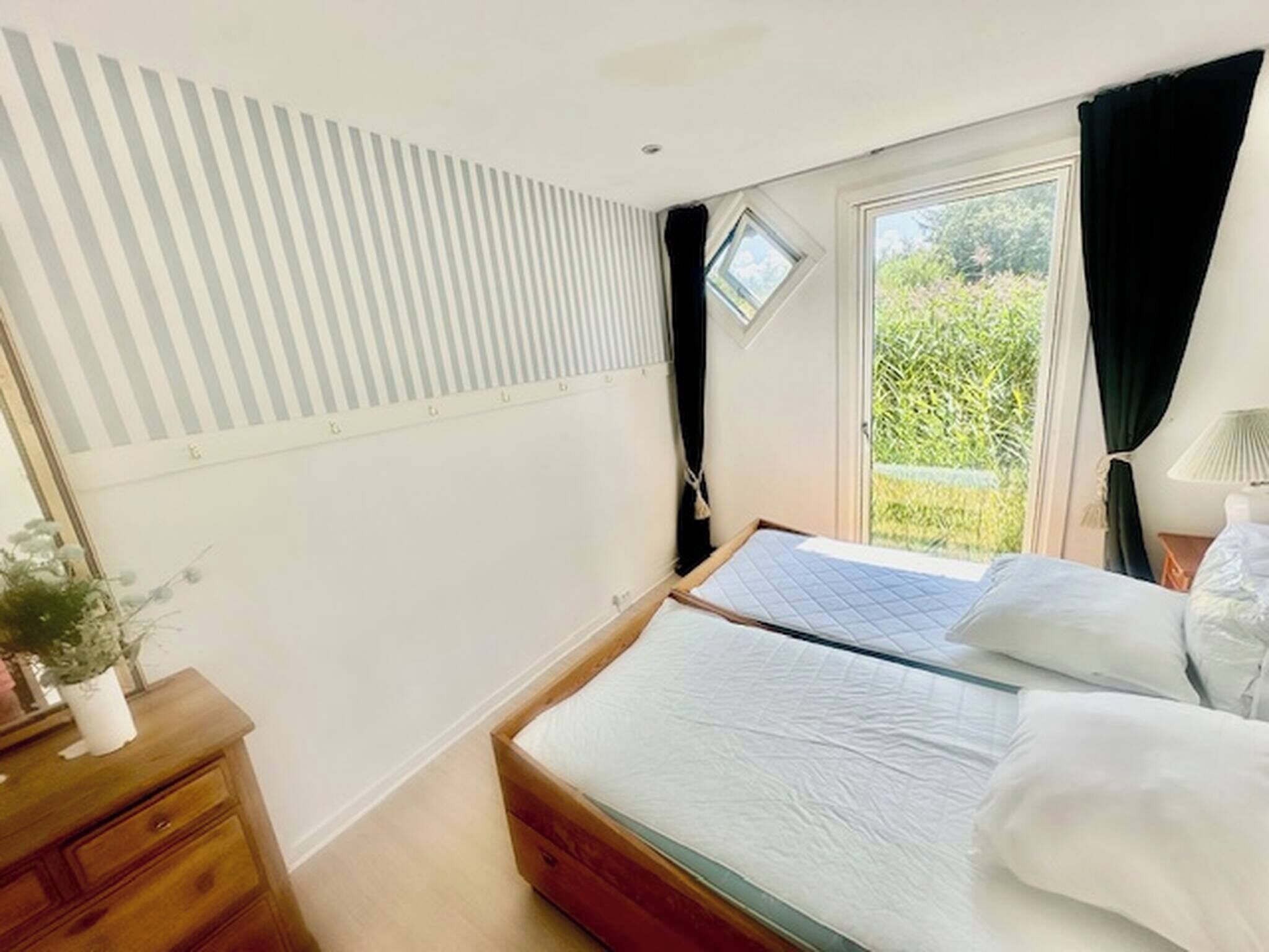 A bedroom with a bed, a nightstand, a potted plant, a window with curtains, and a view of greenery outside.
