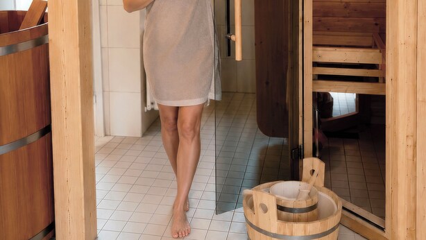 Eine Person mit Handtuch steht in einem Holzsaunaraum mit Glastür, hölzernen Bänken und einer kleinen Holzwanne.