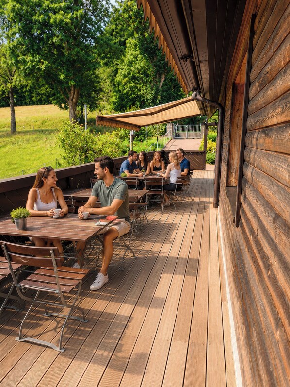 Una terraza de madera con asientos al aire libre, rodeada de follaje y un edificio con techo inclinado.
