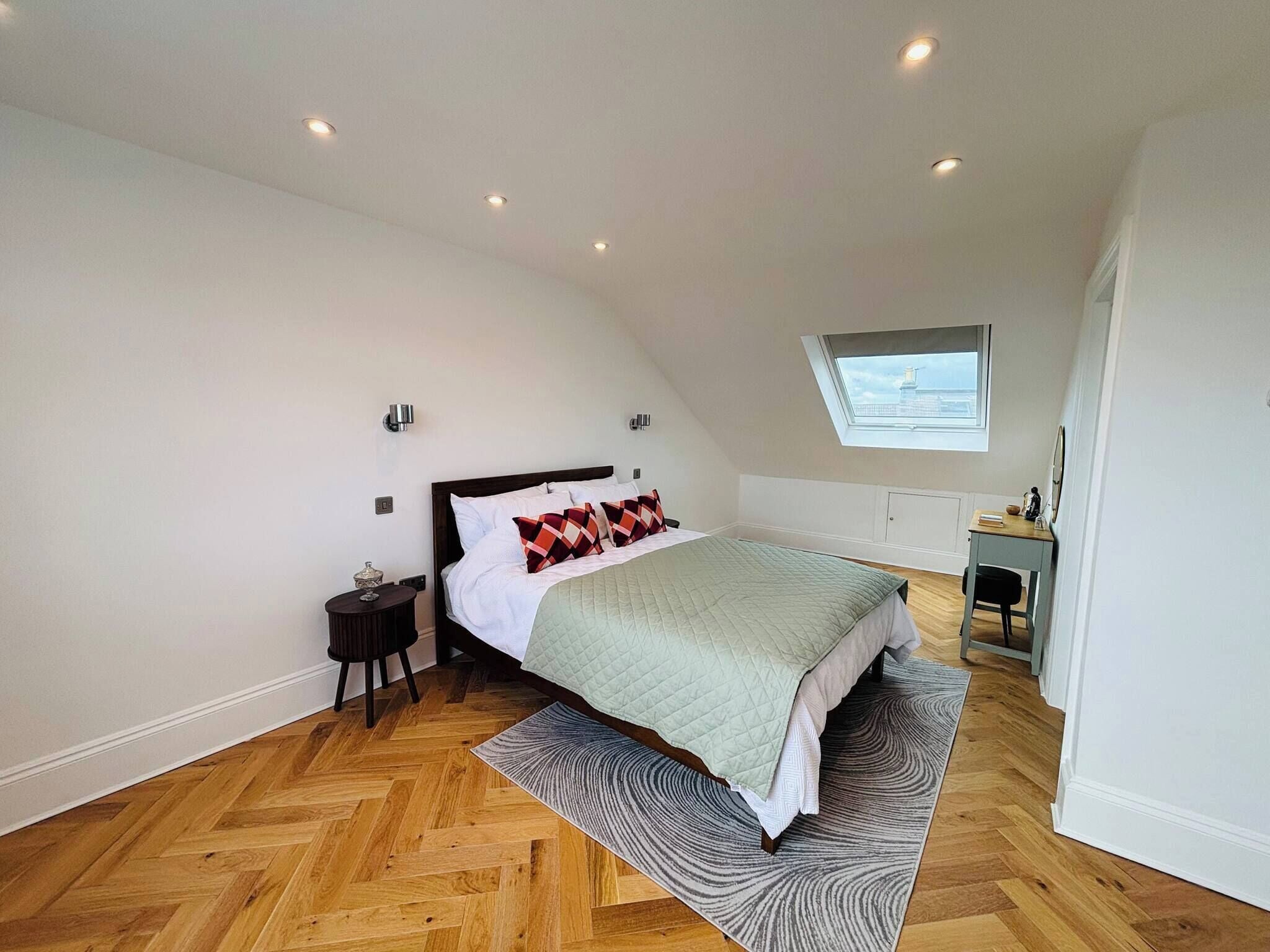 Une chambre avec un plafond en pente, un lit recouvert d’une courtepointe matelassée, une table de chevet, un bureau avec une chaise et un puits de lumière.