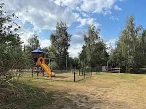 Ein Spielplatz mit einer gelben Rutsche und einer Spielkonstruktion in Rot und Blau, umgeben von Bäumen und einem Zaun.