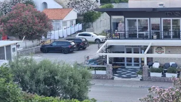 Un edificio con ingresso coperto, un'area di parcheggio con auto e un giardino alberato.