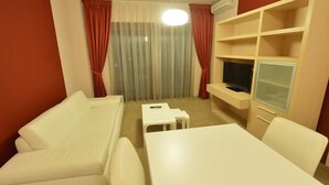 Una stanza piccola e arredata con un divano bianco, un tavolo bianco, una TV su un mobile e una finestra con tende.