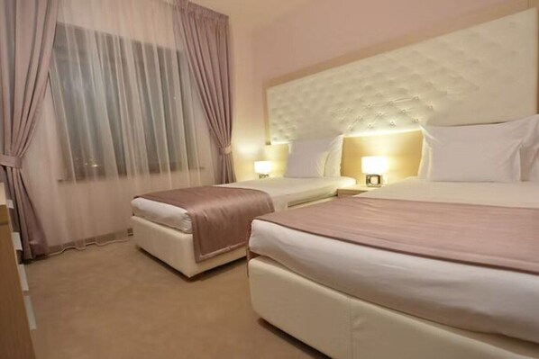A hotel room with two beds, a large padded headboard, and a window with sheer curtains.