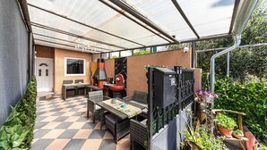 Un patio cubierto con piso a cuadros, mobiliario exterior, una parrilla y plantas en macetas.