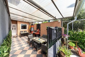 Ein überdachter Patio-Bereich mit Schachbrettmuster-Boden, Gartenmöbeln, einem Grill und Blumentöpfen.