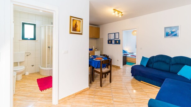A living room with a blue sofa, a dining area with a table set for two, and a kitchen with wooden cabinets. - Apartments Casa Vanna - One Bedroom Apartment (Dubrovnik)