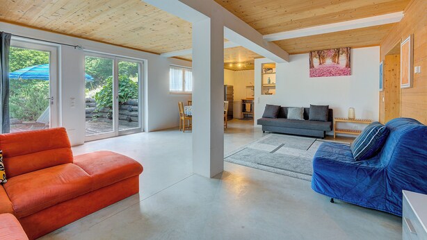 A spacious living room with a large window, a sofa, a blue couch, and a kitchen in the background. - Casa Maamii Mugena With Terrace And Parking (Alto Malcantone)