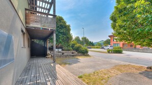 A wooden deck leading to a building with a covered entrance, a parking lot, and a tree-lined street. - Casa Maamii Mugena With Terrace And Parking (Alto Malcantone)