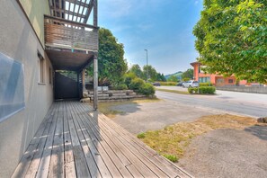 A wooden deck leading to a building with a covered entrance, a parking lot, and a tree-lined street. - Casa Maamii Mugena With Terrace And Parking (Alto Malcantone)