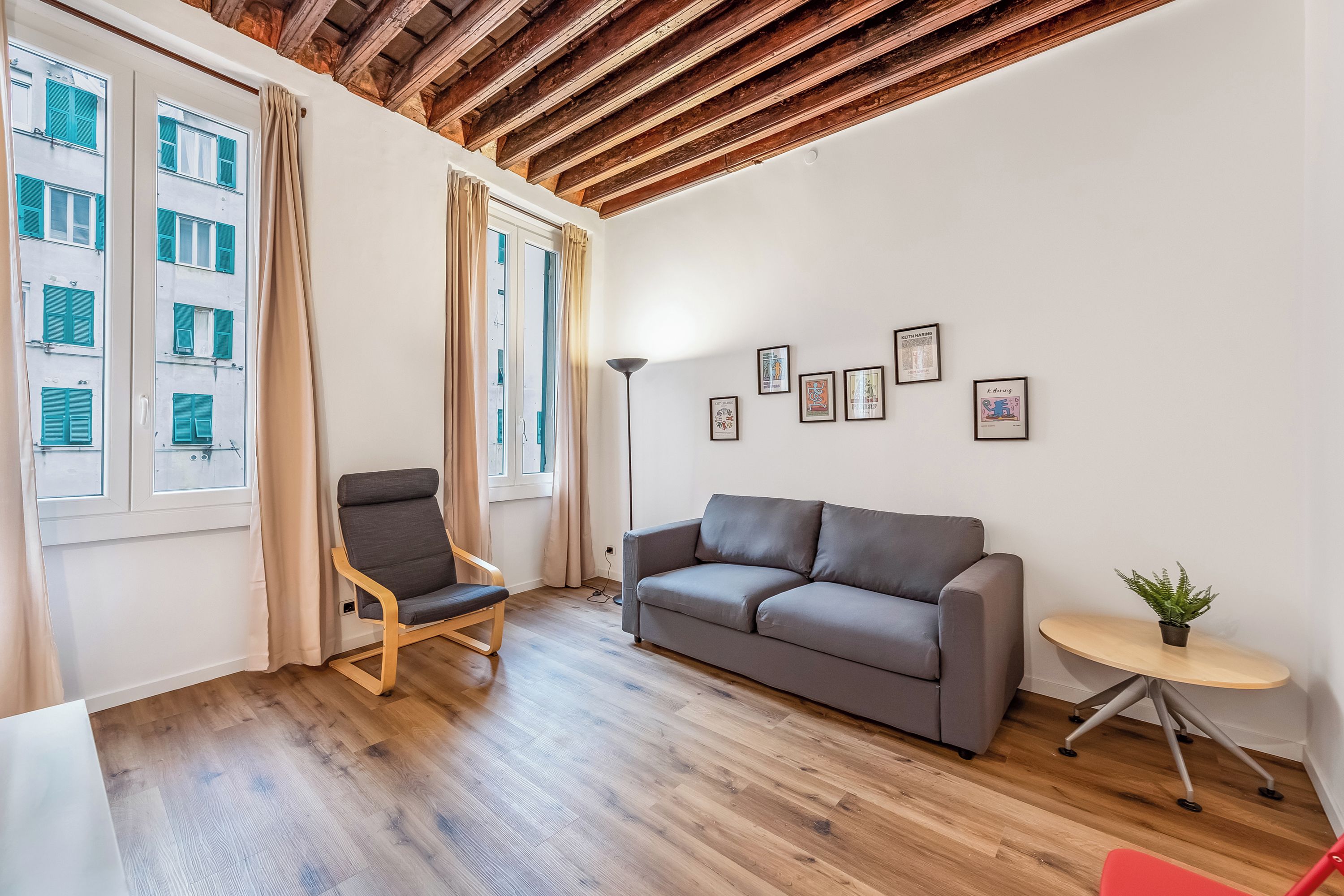 A modern living room with a wooden ceiling, a grey sofa, a wooden chair, a small round table with a plant, and a wall with framed artwork.
