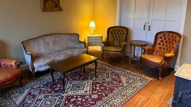 Ein Zimmer mit einer Couch, zwei Sesseln, einem kleinen Tisch, einer Lampe, einem gemusterten Teppich und einer Tür.