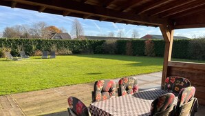 An outdoor patio with a checkered table and floral chairs, a wooden pergola overhead, and a well-maintained lawn in the background.