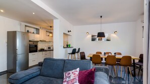 Un soggiorno moderno con un divano grigio, un'area pranzo con tavolo e sedie in legno, e una cucina con mobili e elettrodomestici bianchi.