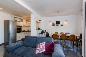 Ein modernes Wohnzimmer mit einem grauen Sofa, einem Essbereich mit Holztisch und -stühlen sowie einer Küche mit weißen Schränken und Geräten.