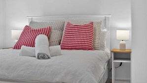Una cama bien hecha con ropa blanca, una toalla enrollada y varias almohadas en rojo y beige. Hay dos lámparas de noche de madera.