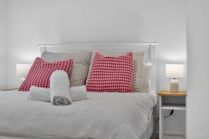 Ein ordentlich bezogenes Bett mit weißer Bettwäsche, ein aufgerolltes Handtuch und mehrere Kissen in Rot und Beige. Auf Holz-Nachttischen stehen je eine Nachttischlampe.