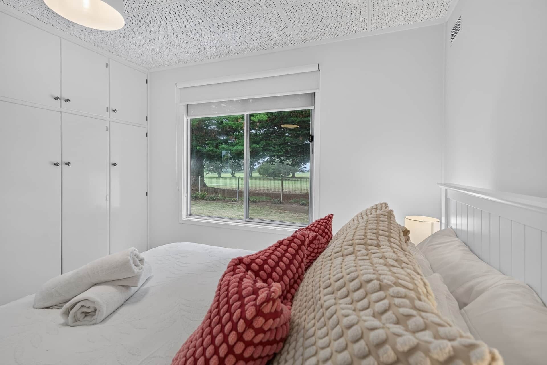 A bedroom with a large window, white walls, and a bed with rolled white towels.