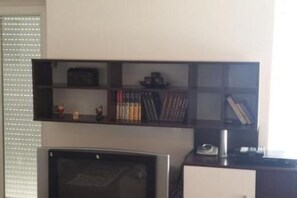 Ein Zimmer mit Fernseher, einem Regal mit Büchern und Dekoration sowie einem Fenster mit Jalousien.