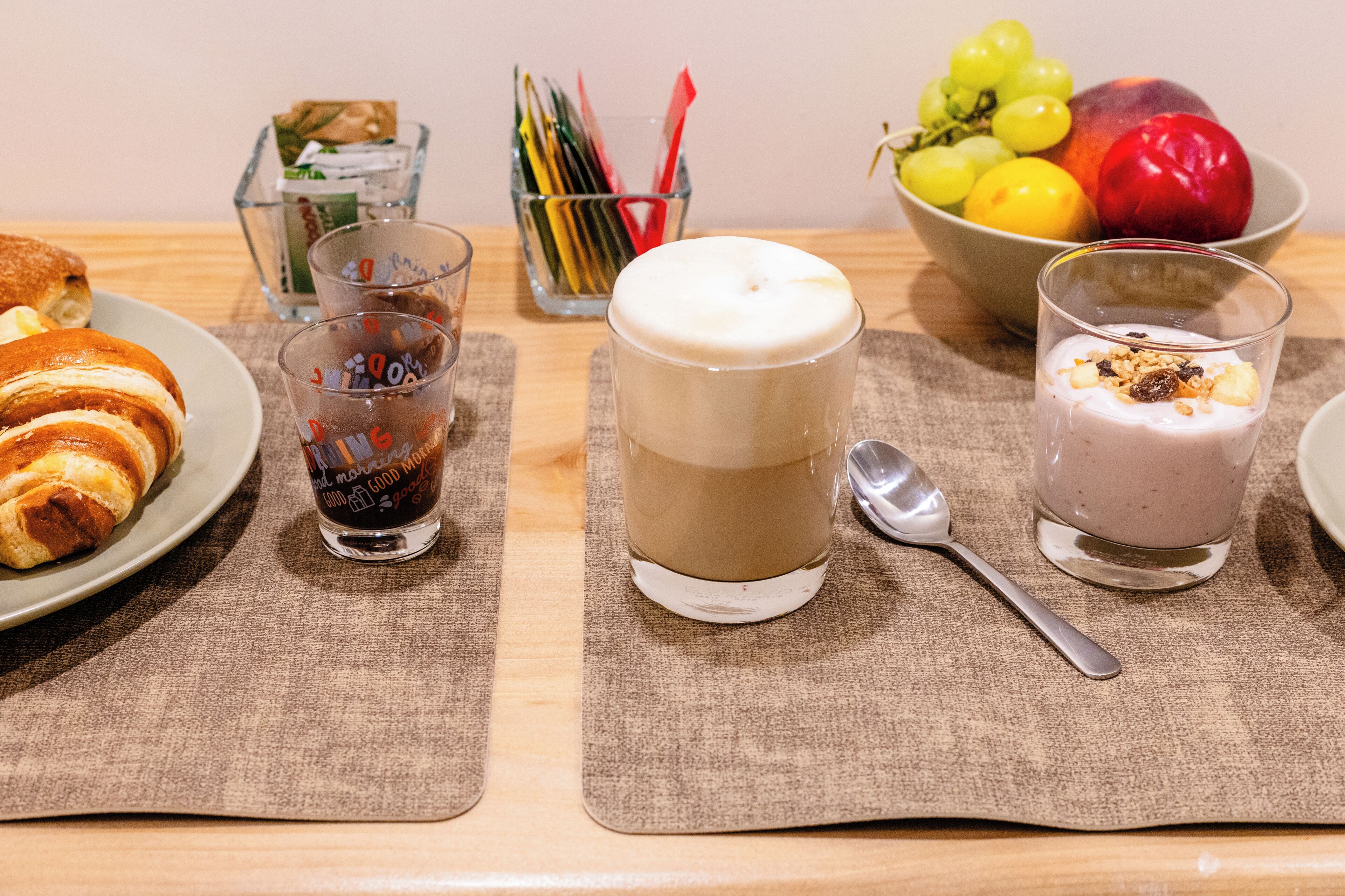 A breakfast table with croissants, a glass of dark liquid, a layered beverage, a bowl of fruit, and a glass of yogurt with granola.