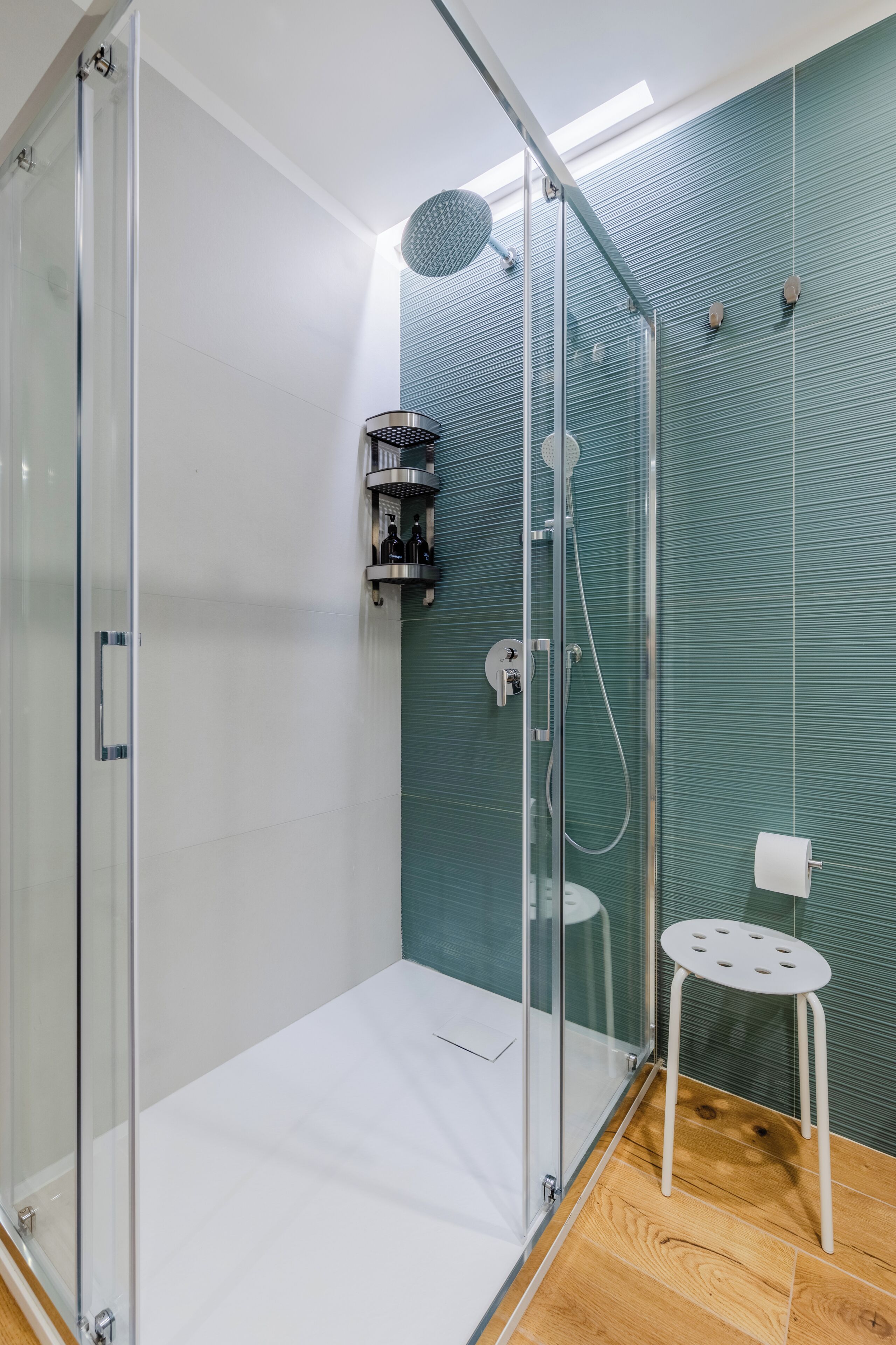 A modern shower area with a glass door, a handheld showerhead, a wall-mounted faucet, and a small white stool.