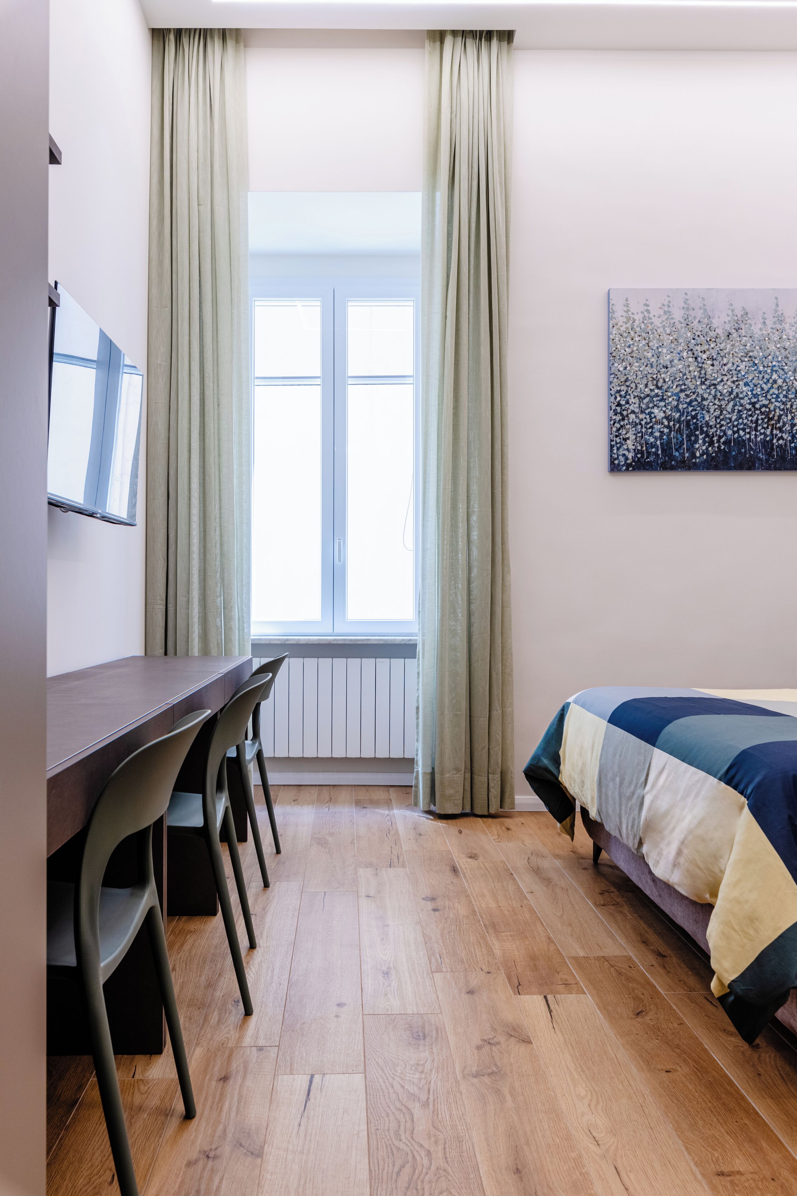 Quarto com piso de madeira, cama com colcha multicolorida, escrivaninha com cadeiras e janela com cortinas.