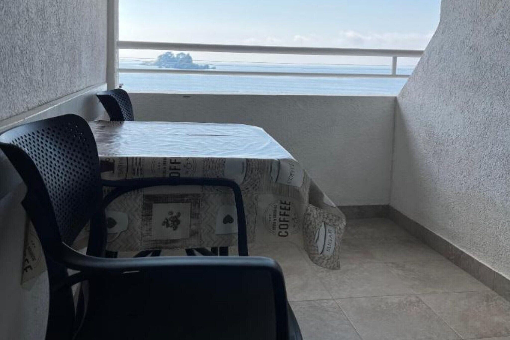 Un petit balcon avec une table recouverte d’une nappe à motifs de café, deux chaises et une vue sur la mer.