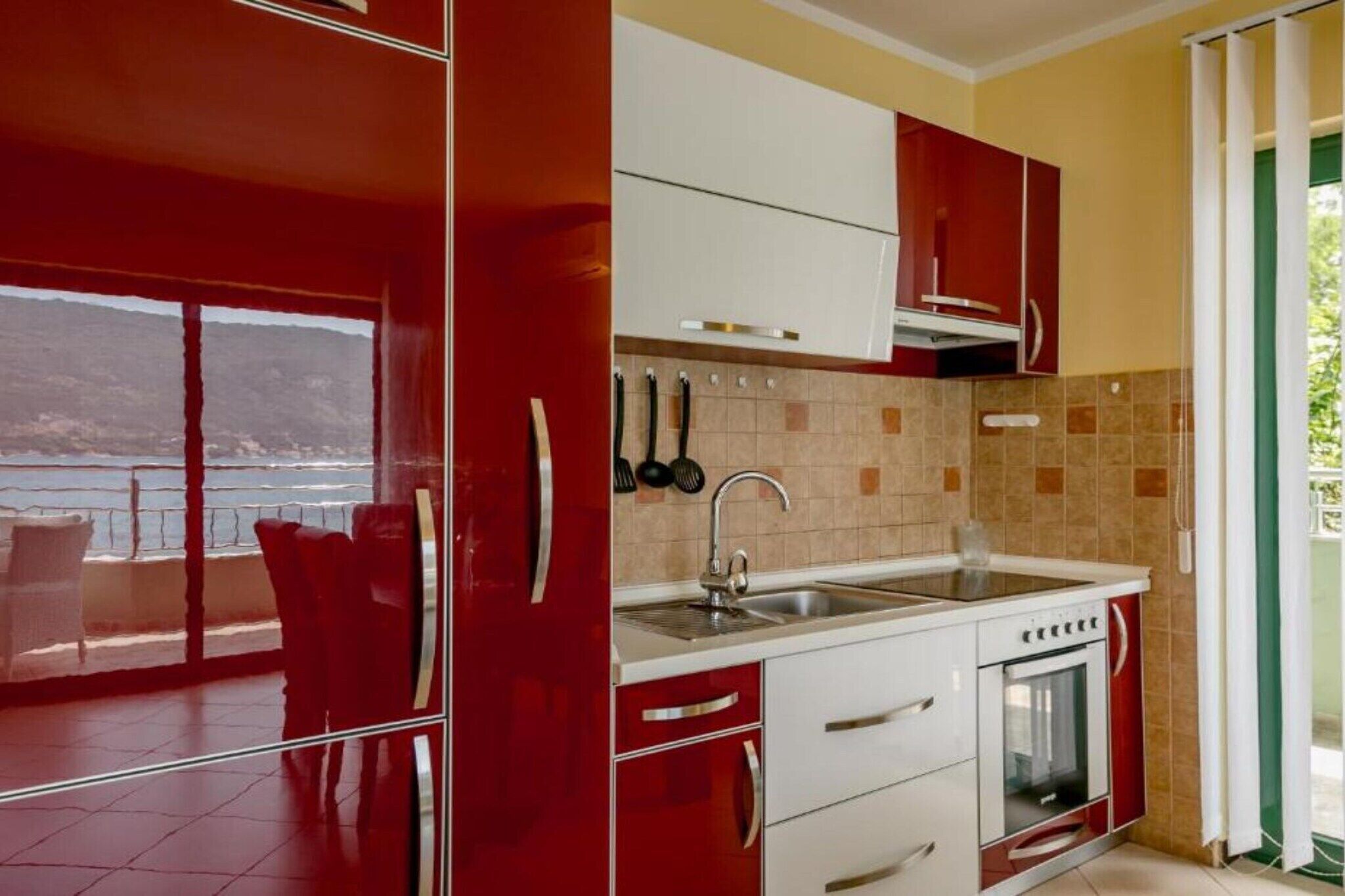 A modern kitchen with red cabinets, a stainless steel sink, and a view of the sea through the sliding glass door.