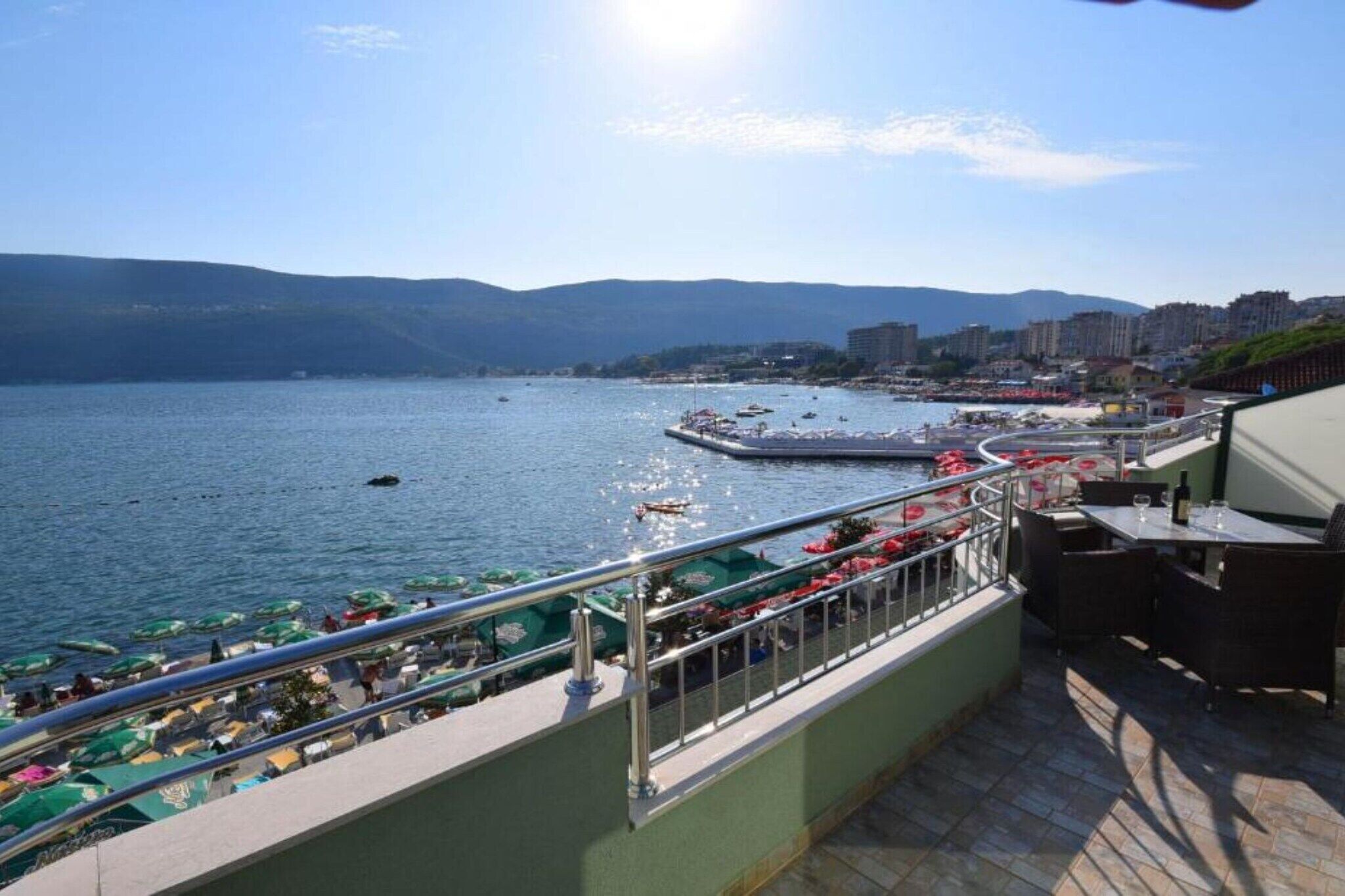 En terrasse ved vandet med udsigt over havet, en bro og fjerne bygninger.