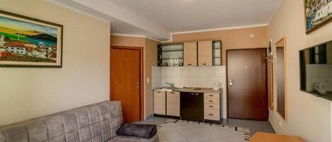 A compact living space with a sofa, a small dining table with fruit, a kitchen area, and a TV on the wall.