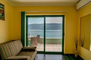 Una habitación con sofá, puerta corrediza de vidrio que da a un balcón con vistas al mar y las colinas.